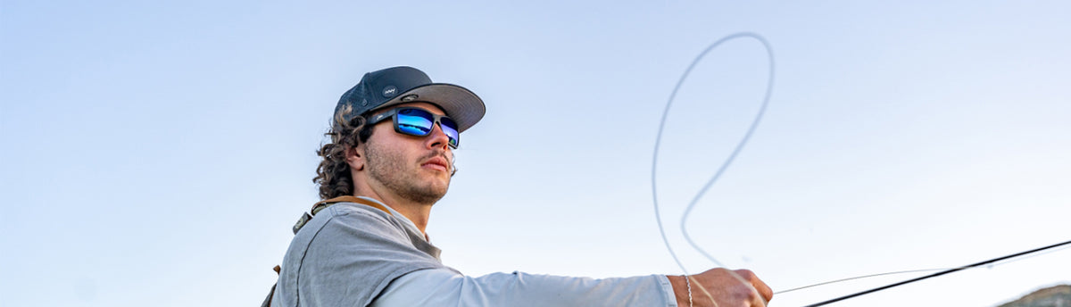 Man fly fishing with sunglasses and a cap against a clear sky