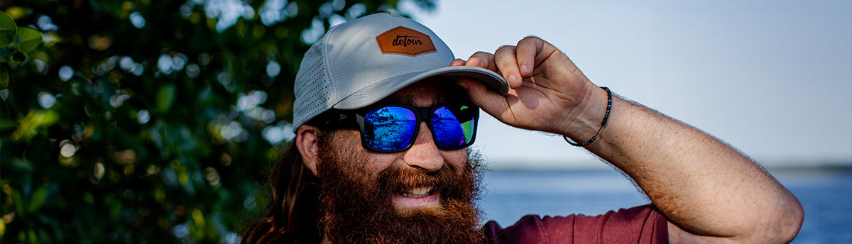 A smiling bearded man wearing a light gray mesh trucker hat with a brown leather patch that reads 'detour', blue mirrored sunglasses, and a burgundy t-shirt, outdoors with green foliage and a blue sky in the background.