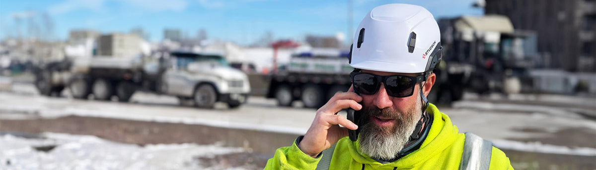 Man in high-visibility jacket and helmet talking on a phone with trucks in the background wearing z87 certified sunglasses
